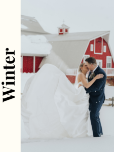 The bride and groom share a romantic kiss in front of a red barn, surrounded by a winter landscape with gently falling snow, creating a magical and intimate moment