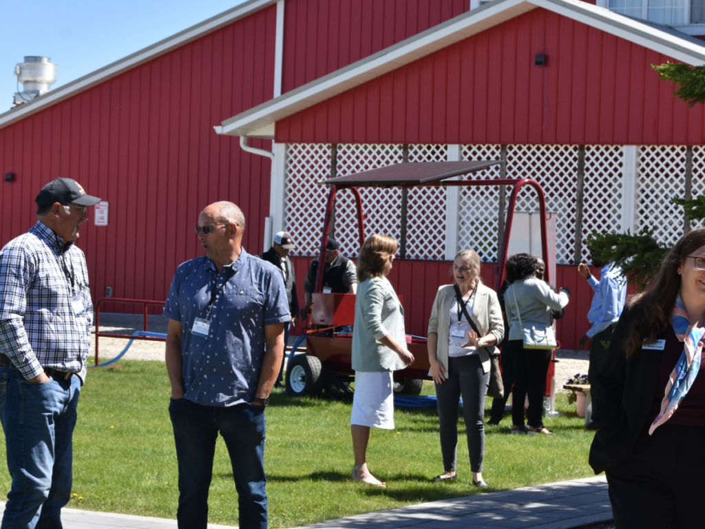 Men and women engaged in discussion near agriculture equipment displayed in front of a barn-style conference venue set in a scenic country setting, with rustic architecture and surrounding greenery enhancing the atmosphere.