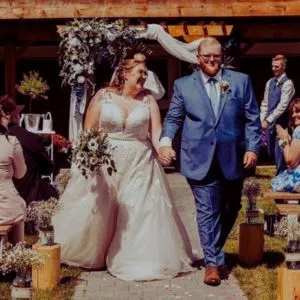 "Bride and groom walking hand in hand down the aisle at an outdoor ceremony at a country wedding venue, surrounded by wooden benches, floral decorations, and a lush natural backdrop