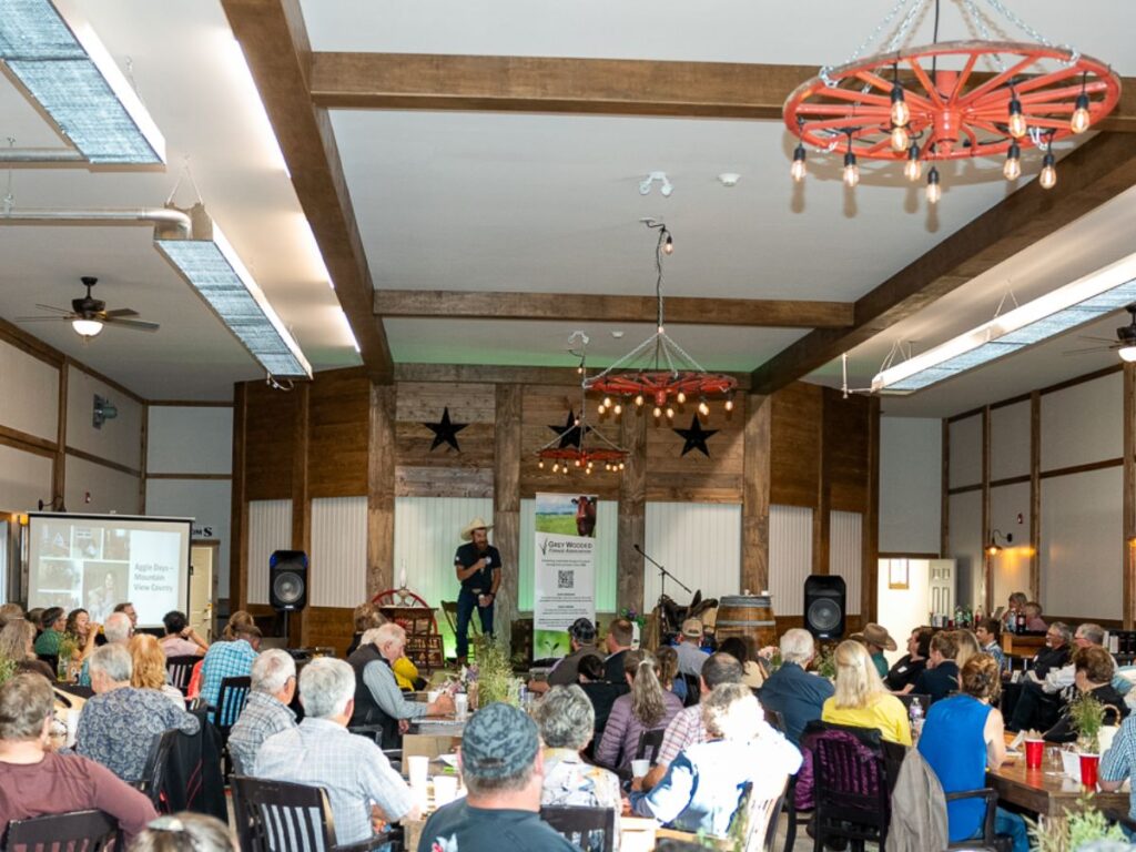 A country event venue near Calgary featuring an entertainer on stage wearing a cowboy hat, delivering comedy to an engaged audience. The room boasts a Western theme with rustic wooden accents, custom chandeliers, and guests seated at tables, enjoying the lively atmosphere.