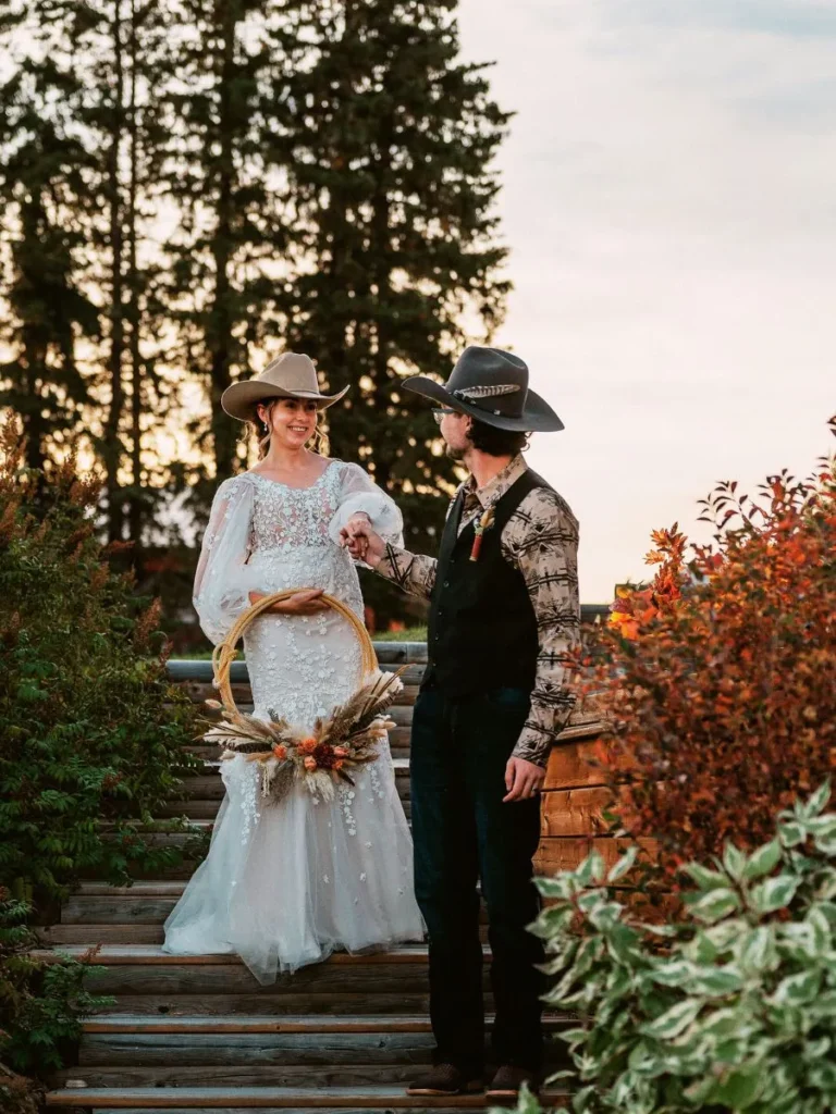 Bride and groom wearing cowboy hats on an outdoor staircase at a rustic country wedding venue, surrounded by picturesque countryside.