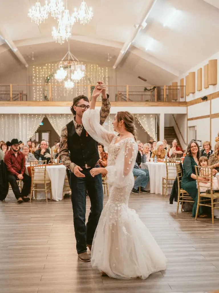 Groom twirling his bride on the dance floor of a stunning country wedding venue with sparkle walls, and a warm ambiance, as friends watch and smile in celebration.