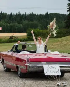 Groom driving a red convertible with the bride standing up, holding her bouquet high, celebrating their joyful wedding ride.