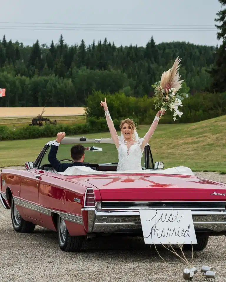 Just-Married Getaway at The Heritage Centre | The Heritage Centre by Mountain View Events Ltd. | The Heritage Centre by Mountain View Events Ltd. Bride raises bouquet from a vintage red convertible with a “Just Married” sign and tin cans, departing The Heritage Centre near Calgary.