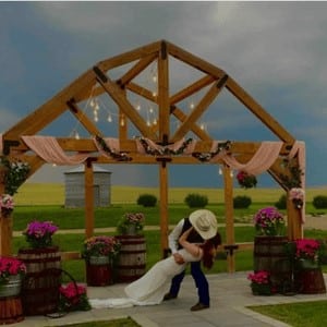 Newlywed groom wearing a cowboy hat kissing his bride under a beautiful wooden arch, surrounded by a scenic outdoor setting