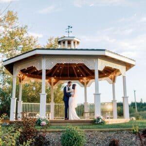 Bride and groom embrace under the white garden gazebo with string lights at The Heritage Centre, an outdoor country wedding venue near Calgary, Alberta.