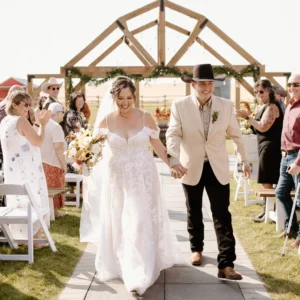 Newlyweds walk down the aisle as guests cheer at an outdoor ceremony beneath a wooden arch.