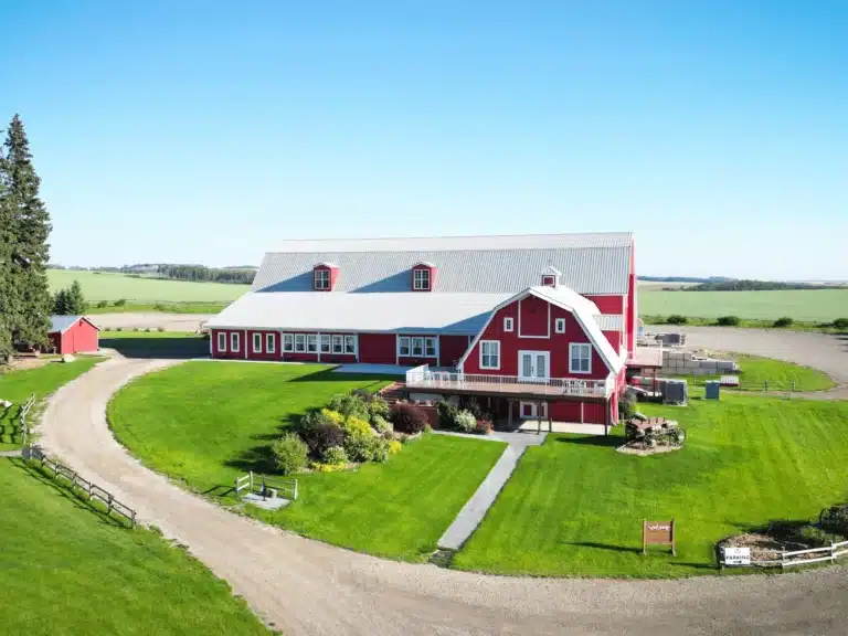 Aerial View of The Heritage Centre | The Heritage Centre by Mountain View Events Ltd. | The Heritage Centre by Mountain View Events Ltd. Aerial view of The Heritage Centre’s red barn venue surrounded by manicured green lawns, gardens, and open prairie landscape under a clear blue sky.