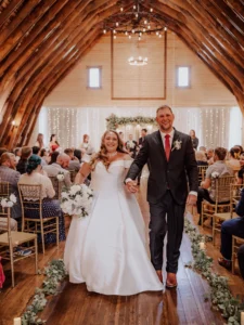 Timber-Arch Ballroom Aisle Moment – The Heritage Centre | The Heritage Centre by Mountain View Events Ltd. | The Heritage Centre by Mountain View Events Ltd. Newlyweds walking down the aisle inside a timber-arched barn with string lights and guests seated.