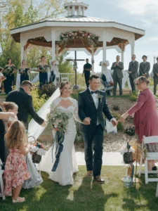 White Gazebo Ceremony Exit – The Heritage Centre | The Heritage Centre by Mountain View Events Ltd. | The Heritage Centre by Mountain View Events Ltd. Elegant Country Wedding Venue Pricing for these Newlyweds walking down the aisle after an outdoor ceremony at a white gazebo with guests cheering.