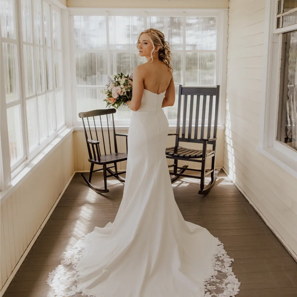 Bride holding a floral bouquet and displaying the lace train of her wedding gown inside the bright enclosed porch of the Heritage Home.