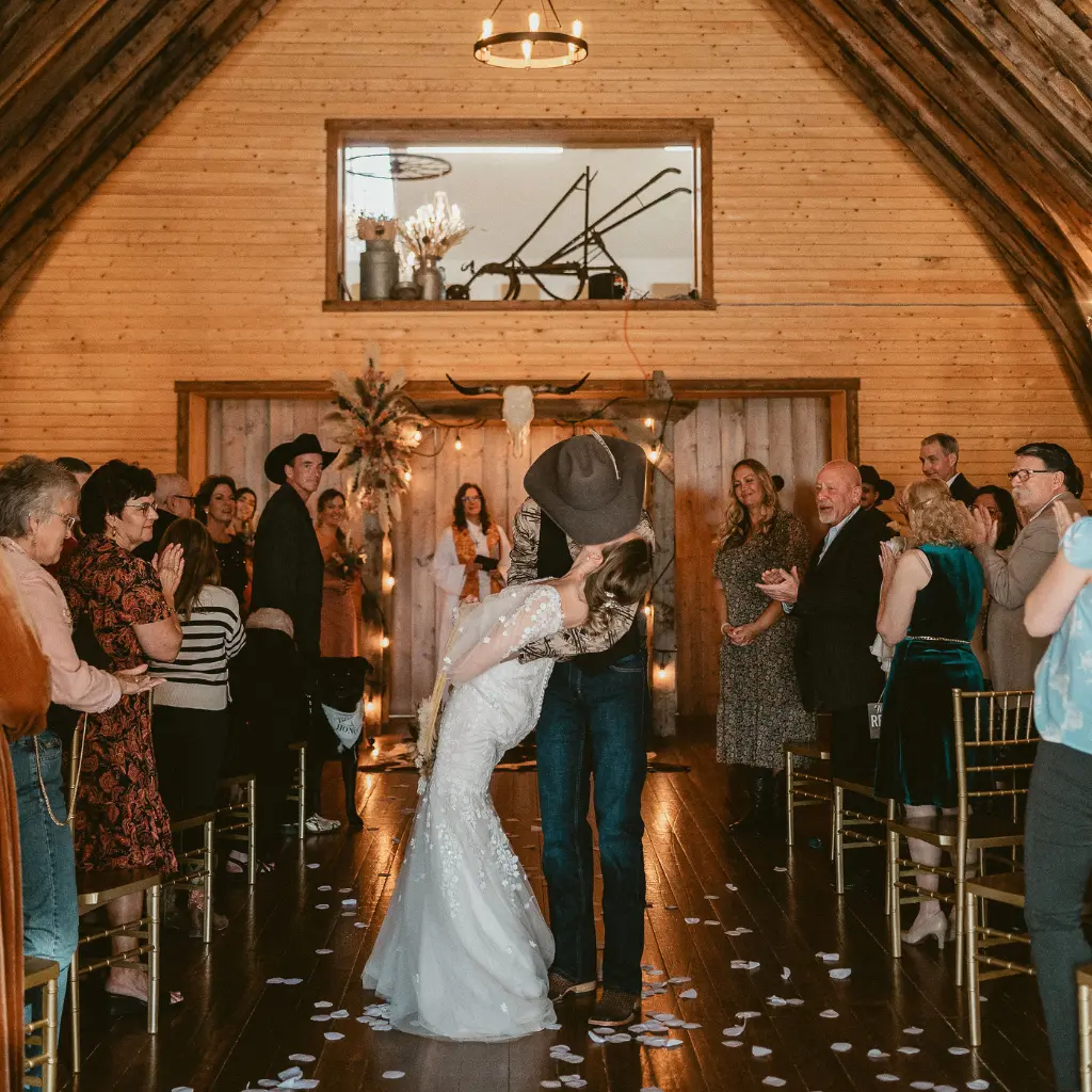 Newlywed couple shares a celebratory kiss while walking down the aisle as guests applaud inside the Historic Barn at The Heritage Centre.