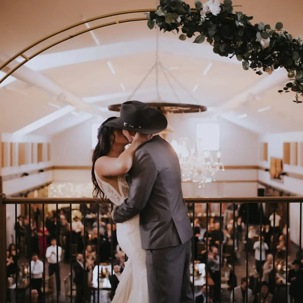 Bride and groom wearing cowboy hats share a kiss on the mezzanine overlooking their wedding reception at The Heritage Centre.