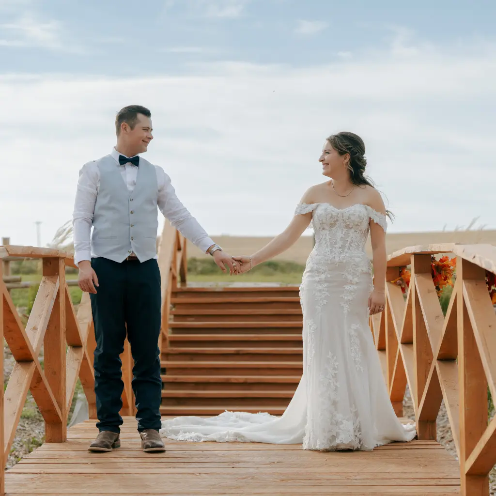 Bride and groom holding hands on a wooden bridge and smiling at each other at The Heritage Centre wedding venue.