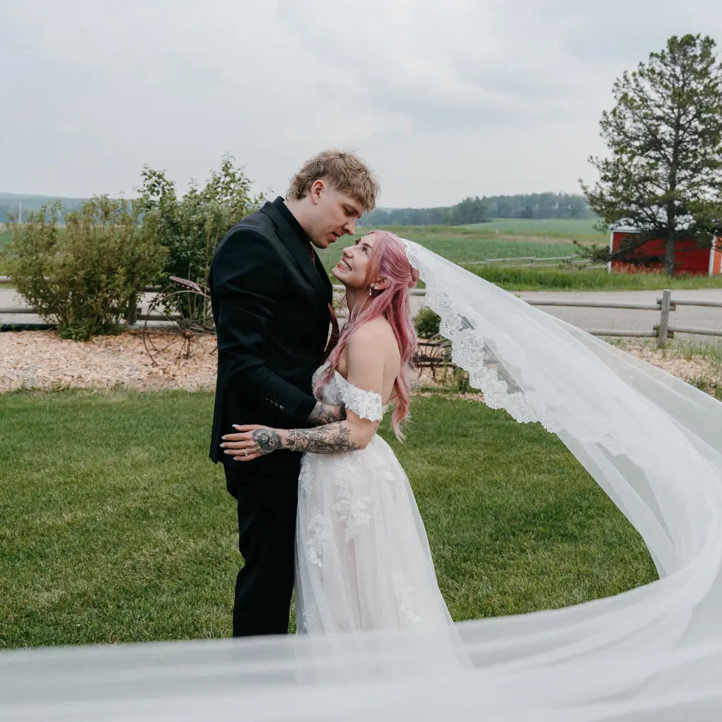 Pink-haired bride and groom embracing on the lawn as her long lace-edged veil flows behind her at The Heritage Centre.
