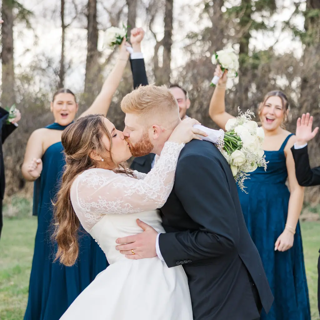 Bride and groom kissing outdoors while their wedding party celebrates behind them at The Heritage Centre.