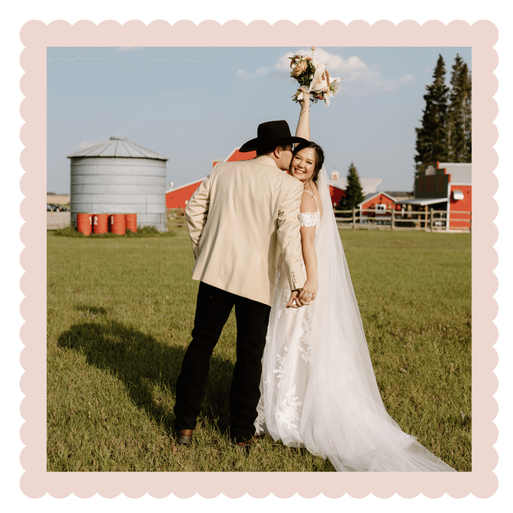 Nicole and Austin hold hands during their outdoor wedding portraits at The Heritage Centre in Cremona, Alberta. The bride smiles while raising her bouquet beside the groom in a cowboy hat, with the venue's iconic red barns and prairie landscape creating a beautiful country wedding backdrop.