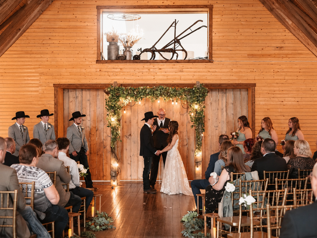 Indoor Wedding Ceremony in the Historic Barn | The Heritage Centre by Mountain View Events Ltd. | The Heritage Centre by Mountain View Events Ltd. Bride and groom exchange vows during an indoor barn wedding ceremony surrounded by greenery, warm lights and gold chairs.