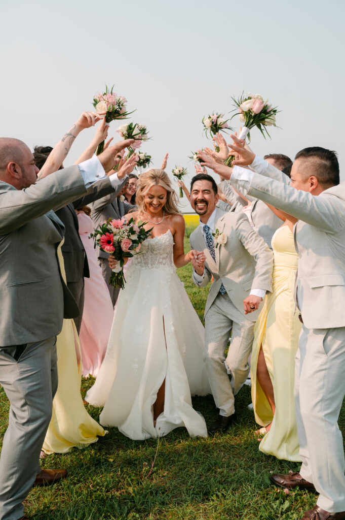 Newlywed couple laughing as their wedding party raises bouquets and creates a celebratory tunnel outdoors at The Heritage Centre.