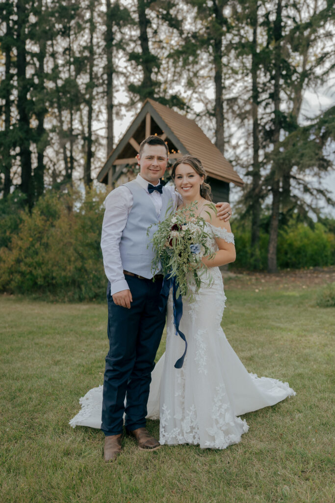 Bride and groom posing together on the lawn in front of a wooden ceremony arch surrounded by tall trees at The Heritage Centre.