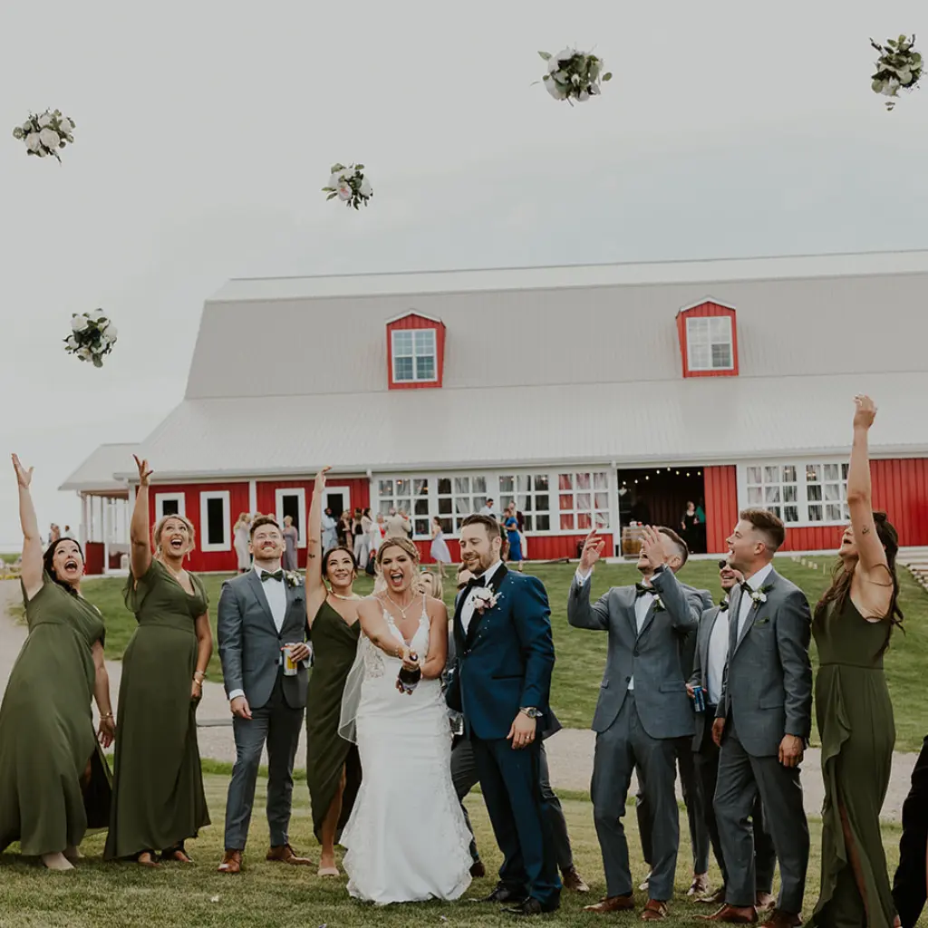 Bride, groom and wedding party laugh as several bouquets are tossed into the air in front of The Heritage Centre’s red barn.