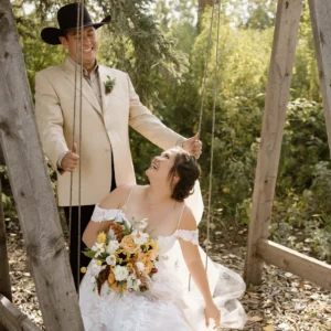 Bride seated on a wooden swing laughing with the groom beside her in a wooded photo area at The Heritage Centre.