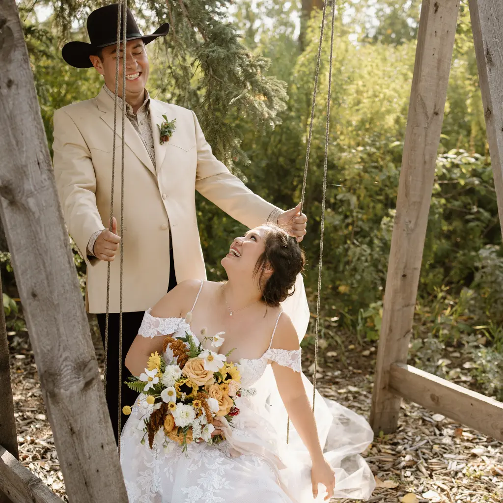 Bride seated on a wooden swing laughing with the groom beside her in a wooded photo area at The Heritage Centre.