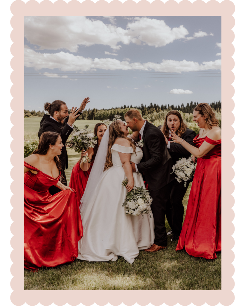 Bride and groom kiss as bridesmaids in red gowns cheer outdoors on a sunny day with bouquets nearby.