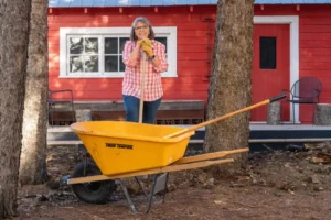 Debora Rice-Salomons smiling and leaning on a shovel beside a wheelbarrow in front of a red building at The Heritage Centre