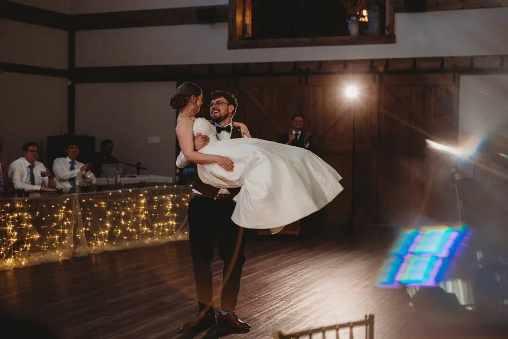 Groom lifting bride in a white wedding gown during their first dance in a rustic barn, with string lights along the head table and guests clapping in the background.