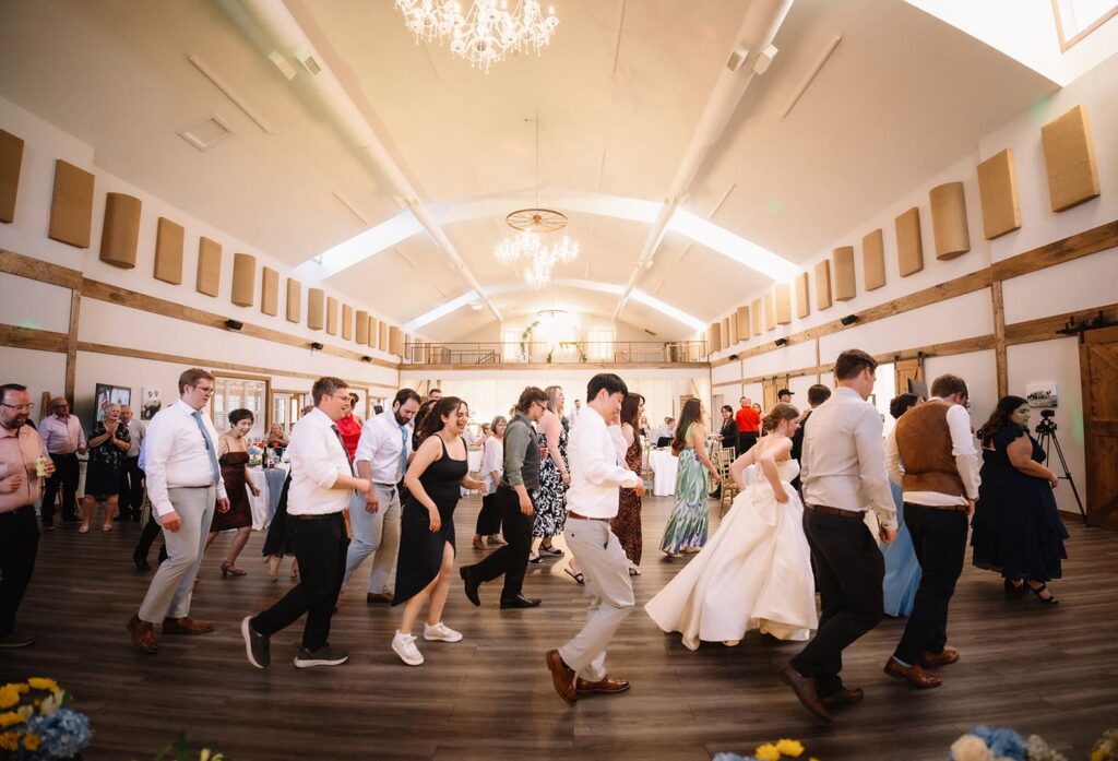 Wedding reception with guests in formal wear dancing on a wooden floor under chandeliers in a bright hall.