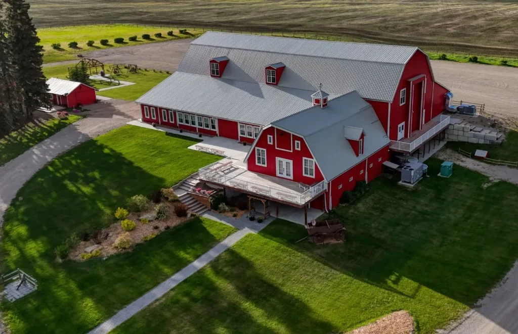 Aerial view of a large red barn with white trim and attached buildings on a manicured green lawn, with gravel driveways and surrounding farmland.