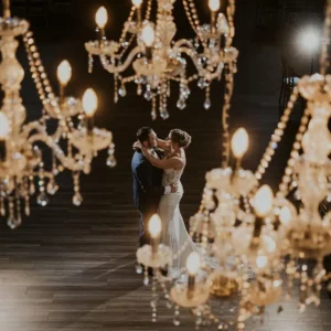 Groom holding his bride in a romantic embrace on the dancefloor, beneath stunning custom chandeliers, during their wedding celebration.